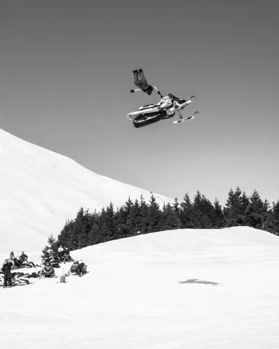 A black and white photo of a snowmobiler performing a high-altitude jump against a mountain backdrop.